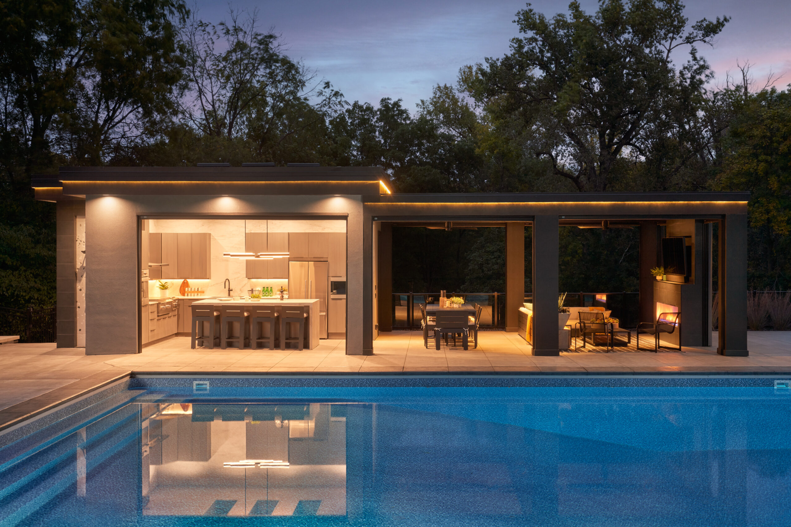 Outdoor in-ground pool with contemporary pool house featuring a bar/kitchen area and sitting area. Night shot.